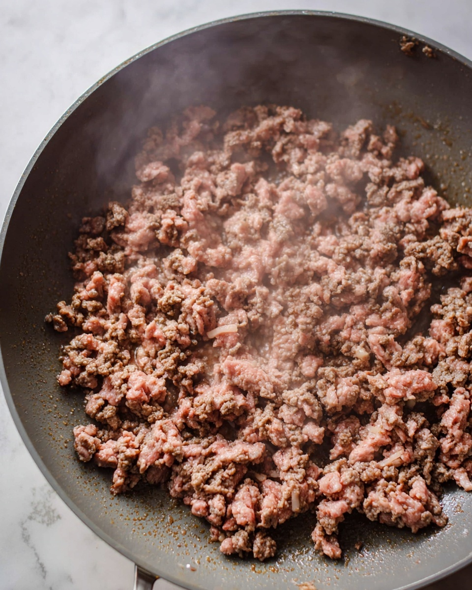 The image shows browned ground meat cooking in a large gray pan. The meat is spread evenly over the pan's surface, showing small, crumbly pieces in a mix of light and dark brown colors with some hints of pink. The pan is on a white marbled surface, and steam rises gently from the cooking meat, highlighting its moist texture and slight gloss. The scene captures the moment when the meat is nearly fully cooked but still has some soft spots in the center. photo taken with an iphone --ar 4:5 --v 7