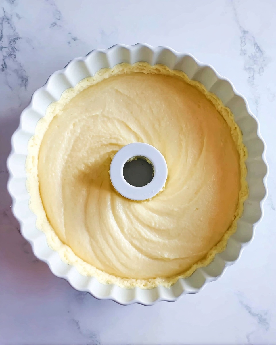 A smooth light yellow batter layer evenly spread inside a fluted white bundt pan with a central tube in the middle; the batter surface is slightly textured with soft swirls and is level with the edges of the pan, all placed on a white marbled surface. photo taken with an iphone --ar 4:5 --v 7