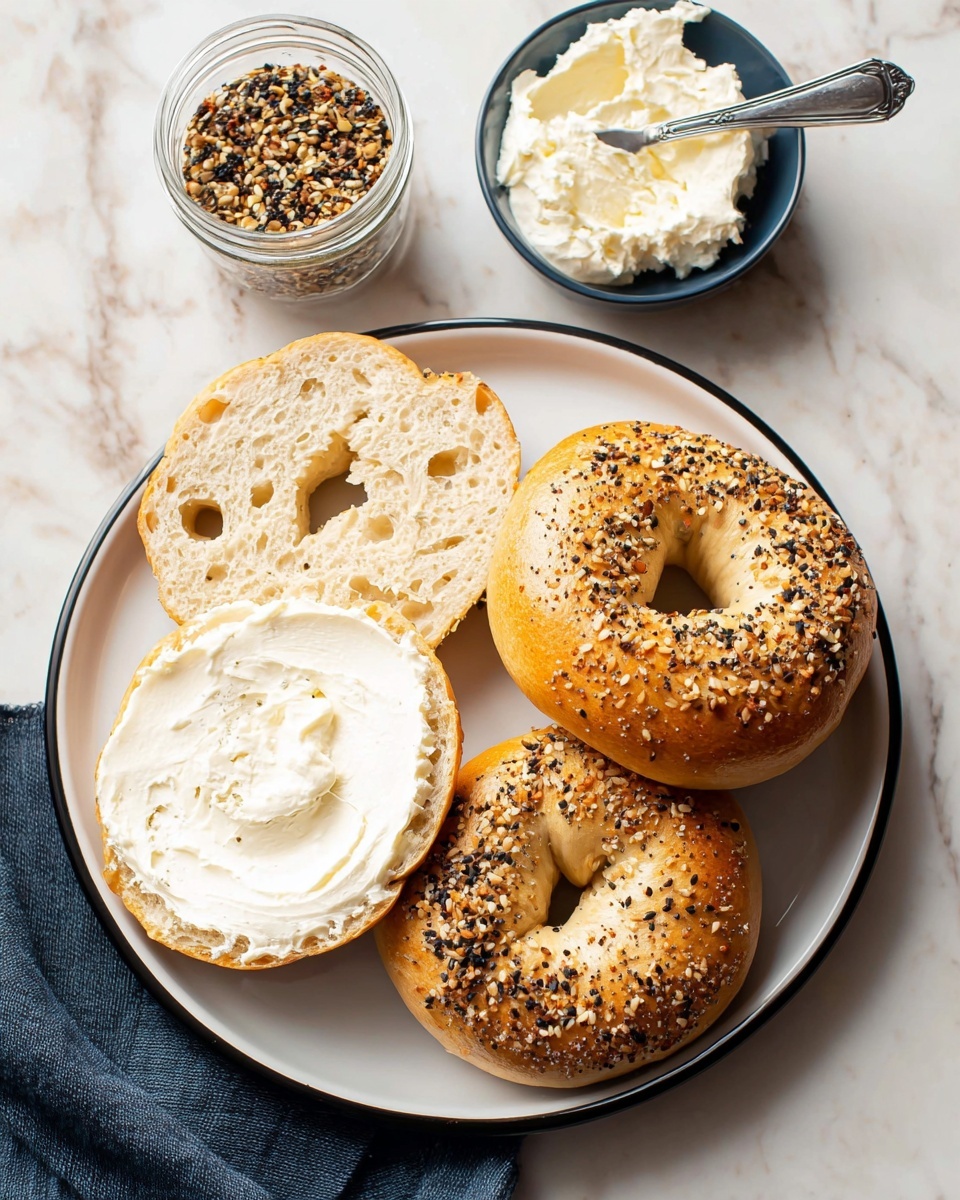 A close-up view of one half of a light beige bagel held by a woman's hand with a gold ring on the ring finger, showing a soft, airy, and slightly porous texture with small holes scattered throughout. The bagel is toasted lightly on the edges with a few tiny black seeds visible inside the crumb. In the blurred background, a white plate holds more whole, round bagels, all on a white marbled surface. Photo taken with an iphone --ar 4:5 --v 7