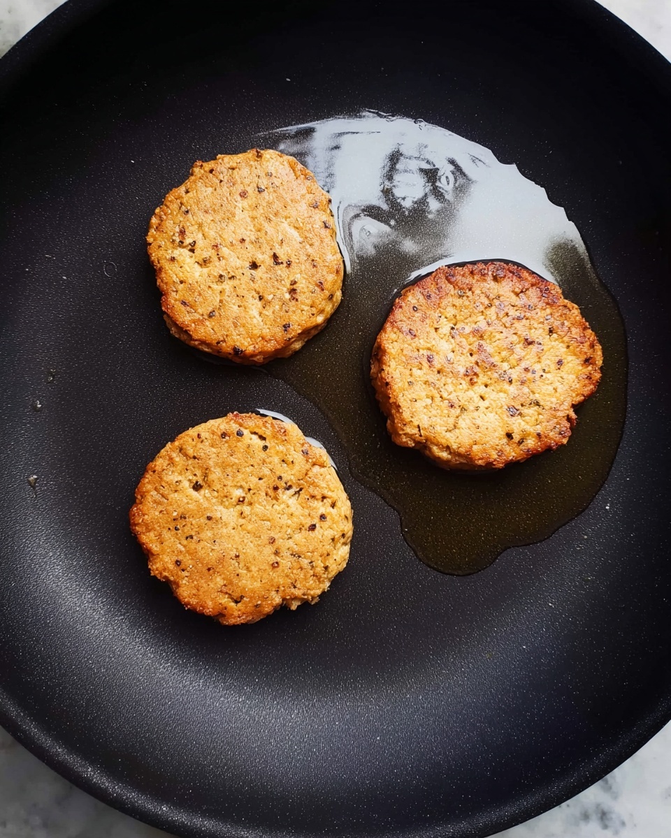 Three light brown, round patties with small black flecks cook in a black pan with a thin layer of oil. The patties have a rough, slightly uneven texture and crispy edges. The pan's matte black surface contrasts with the shiny oil spots around the patties, highlighting their golden color. The scene is simple, focused on the golden patties warming in the pan on a white marbled texture. photo taken with an iphone --ar 4:5 --v 7