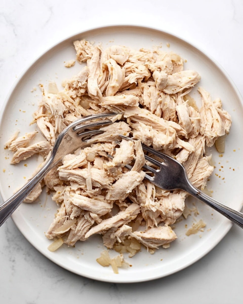 The image shows a white round plate filled with shredded light beige cooked chicken pieces spread evenly across the plate. Two silver forks rest on top of the chicken, one positioned slightly left of center and the other to the right, both angled inward and partially stuck into the chicken. Small bits of translucent cooked onion are scattered around the edges on the white plate. The plate sits on a white marbled surface. Photo taken with an iphone --ar 4:5 --v 7