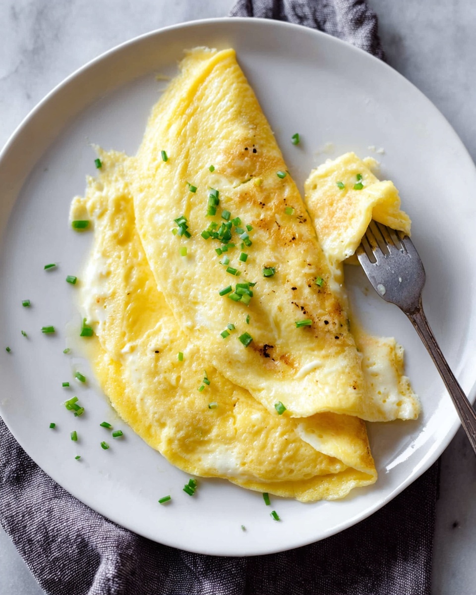 A white plate with a soft, folded yellow omelette that shows a smooth texture with light browning in some spots. Small green chive pieces are sprinkled on top, adding a fresh touch of color. A fork lifts a folded piece of the omelette, revealing a slightly creamy inside. The plate sits on a white marbled surface with a gray cloth nearby. Photo taken with an iphone --ar 4:5 --v 7