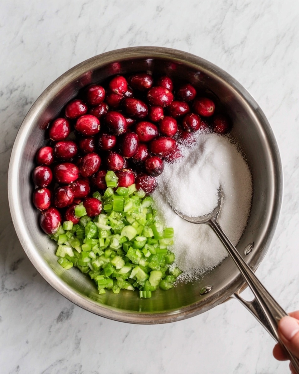 A shiny metal pan holds the ingredients for a recipe, with three distinct sections visible. On the left side, there are whole red cranberries, their smooth round surfaces catching the light. On the right side, there is a heap of fine white sugar, looking powdery and soft. In front of the sugar and cranberries, there are small diced pieces of green celery, fresh and crisp in appearance. The pan sits on a white marbled textured surface. A woman's hand holds a silver spoon resting in the pan. photo taken with an iphone --ar 4:5 --v 7