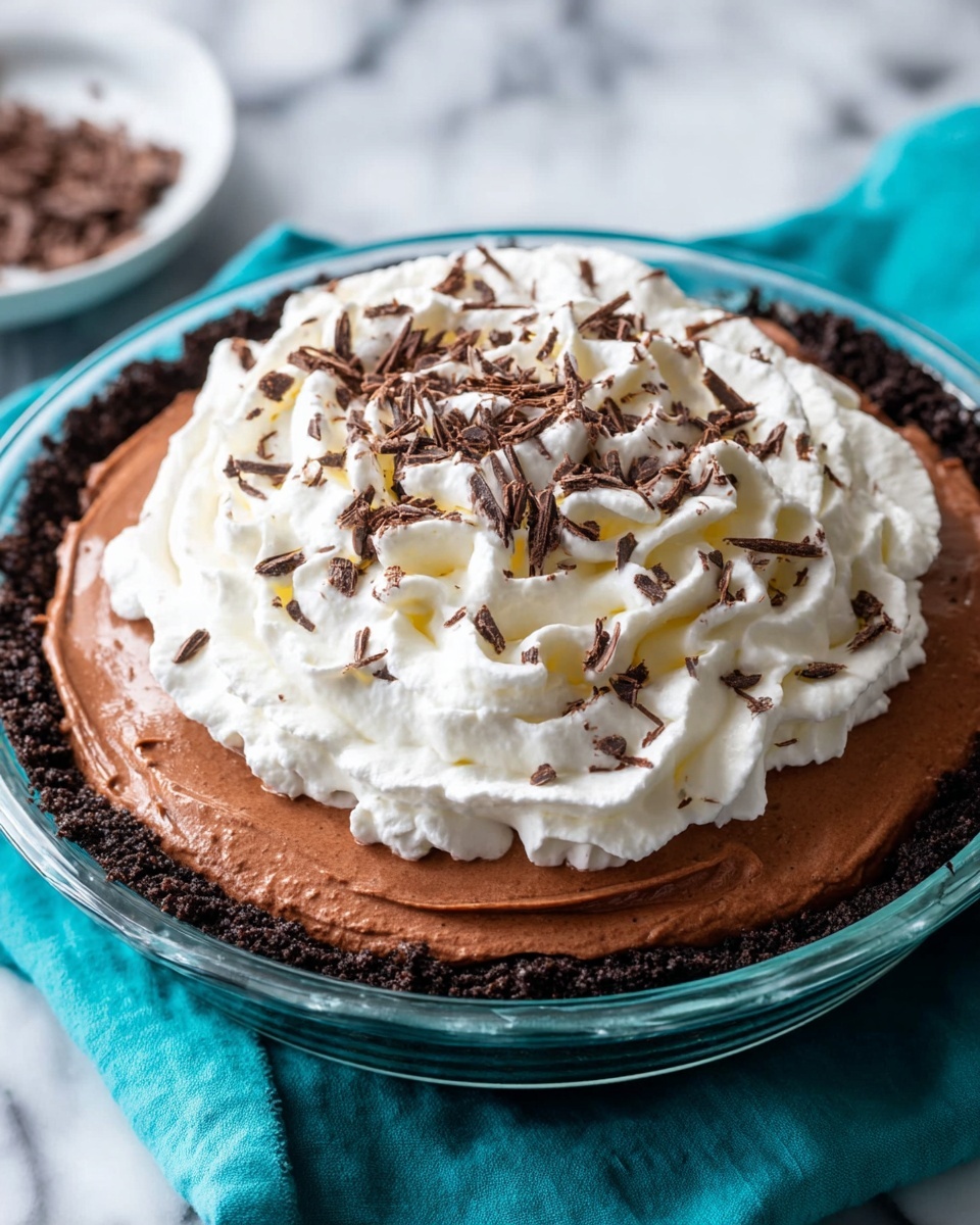 This is a three-layer dessert served in a clear glass pie dish, placed on a white marbled surface with a teal cloth around it. The bottom layer is a dark chocolate crumb crust that forms the base and edges. Above this is a thick, smooth, light brown chocolate filling layer spread evenly towards the crust edges. The top layer is a fluffy, white whipped cream spiral piled high in the center with a soft texture, decorated with small shavings of dark chocolate sprinkled over it. Photo taken with an iphone --ar 4:5 --v 7