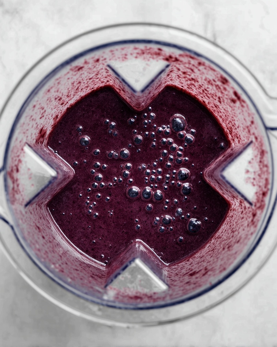 A top-down view of a blender filled with a thick, dark purple smoothie with visible small bubbles on the surface. The blender container is transparent and has a geometric shape with indents. The smoothie fills the blender nearly to the top edge. The surface below the blender is a white marbled texture. photo taken with an iphone --ar 4:5 --v 7