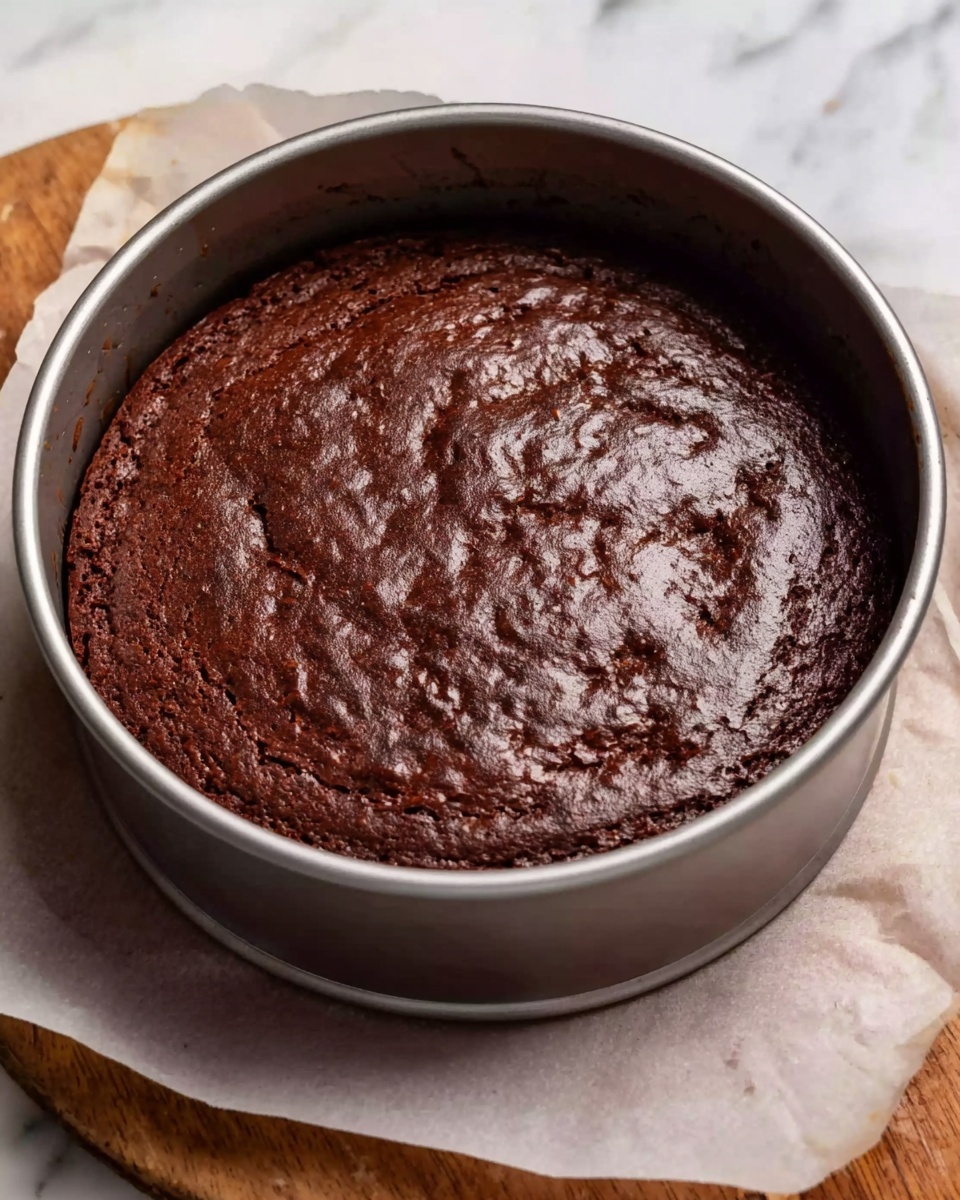 The image shows a freshly baked chocolate cake inside a round metal springform pan placed on white parchment paper. The cake has one layer, with a rich dark brown color and a shiny, slightly cracked surface texture. The pan rests on a wooden surface with a white marbled background visible around it. photo taken with an iphone --ar 4:5 --v 7