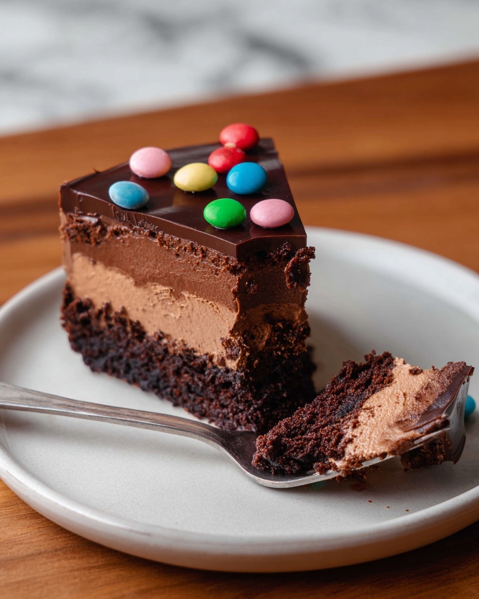 A slice of chocolate cake with three distinct layers sits on a white plate. The bottom layer is dark and dense, resembling a brownie texture. Above it is a thick, lighter brown mousse layer, soft and creamy. The top layer is a smooth, glossy dark chocolate ganache, decorated with small, round, colorful candy pieces in red, green, blue, pink, and orange scattered unevenly. A piece of the cake is held on a silver fork placed on the plate, showing the contrast of the soft mousse and dense brownie. The setting features a wooden table and a white marbled background. photo taken with an iphone --ar 4:5 --v 7