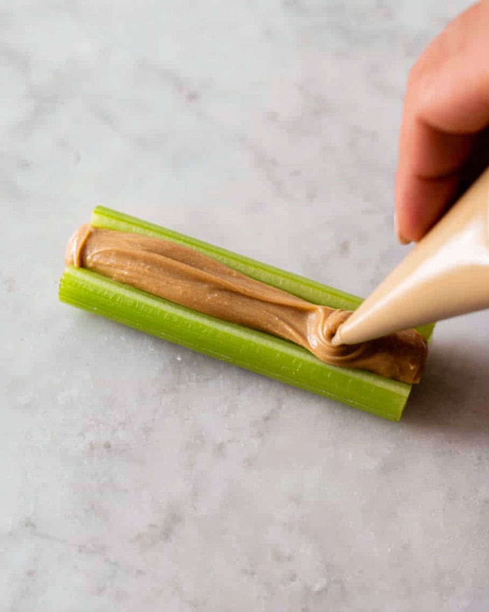 A close-up image shows a woman's hand holding a beige piping bag, squeezing a smooth and creamy light brown spread in a thick line down the center of a fresh green celery stick. The celery is flat on a white marbled surface. The contrast between the bright green celery and the light brown spread is clear and vivid. Photo taken with an iphone --ar 4:5 --v 7