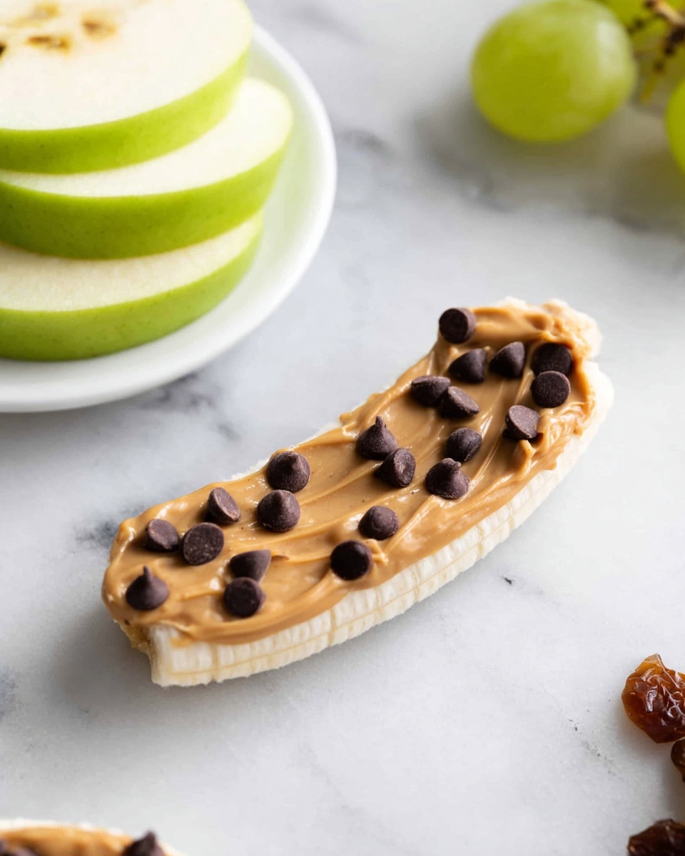A white banana piece lies flat on a white marbled surface, topped with a thick layer of smooth light brown peanut butter, which is sprinkled with small dark brown chocolate chips evenly spread across. In the background, there is a white plate holding two green apple slices layered with peanut butter and raisins in between. Another similar snack with grapes and apple slices is partially visible at the top right corner, all set on the white marbled surface. photo taken with an iphone --ar 4:5 --v 7