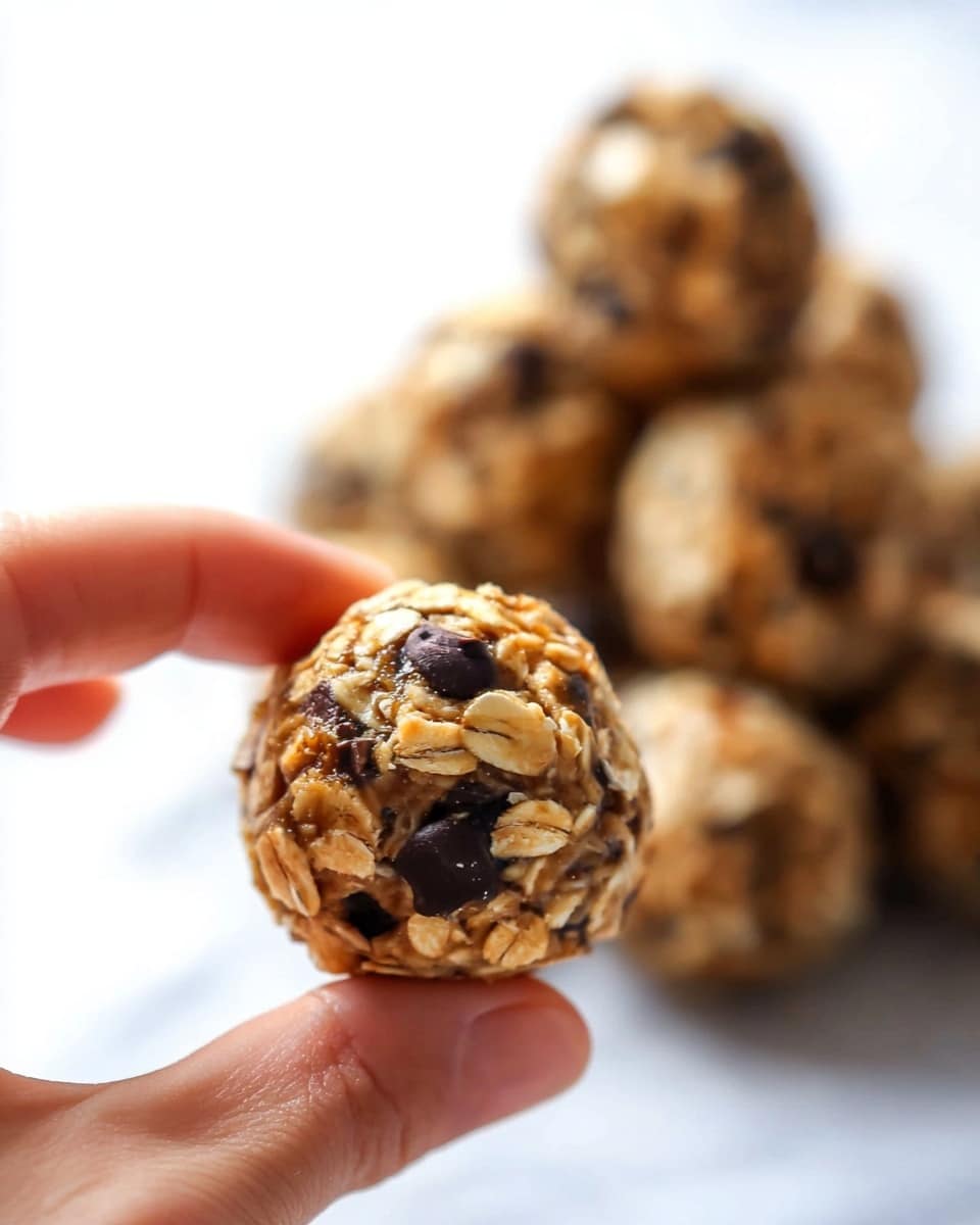 A woman's hand is holding a small round oatmeal ball with visible oats and dark chocolate chips mixed inside. The ball has a rough, textured surface with a mix of light brown and dark brown colors from the oats and chocolate. In the blurry background, there is a pile of similar oatmeal balls, giving a sense of a group of treats stacked together. The setting has a clean white marbled texture beneath the hand and treats. Photo taken with an iphone --ar 4:5 --v 7