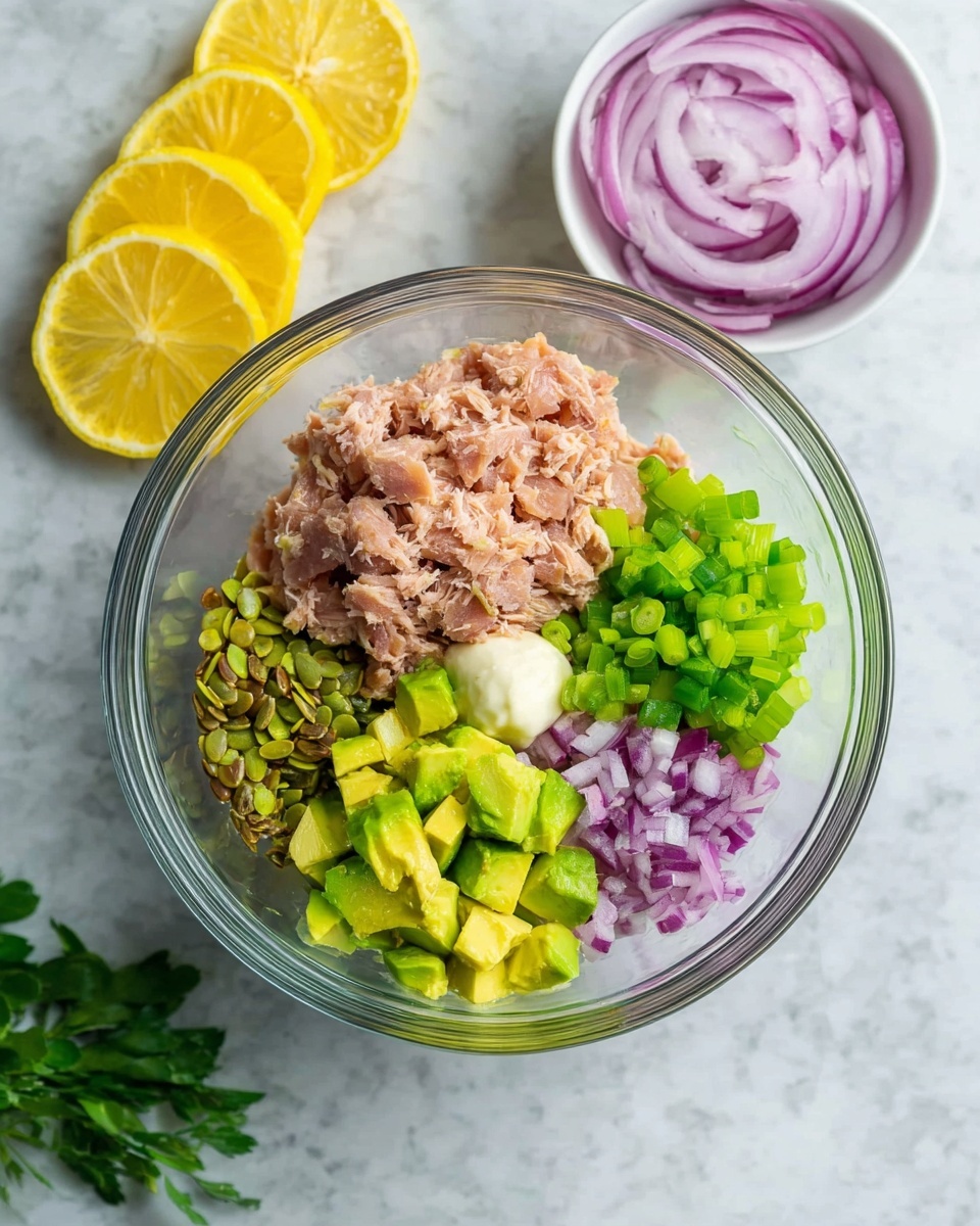 A clear glass bowl contains five separate layers of ingredients arranged inside it: light brown flaky tuna in one section, bright green diced avocado in another, small white mayo dollop next to the avocado, finely chopped purple onion pieces in a small pile, and light green chopped celery with some mixed seeds on top. Nearby, there is a small white bowl filled with thinly sliced red onion rings and two slices of grilled lemon, all set on a white marbled surface. Some green herb leaves are placed around the bowl. Photo taken with an iphone --ar 4:5 --v 7