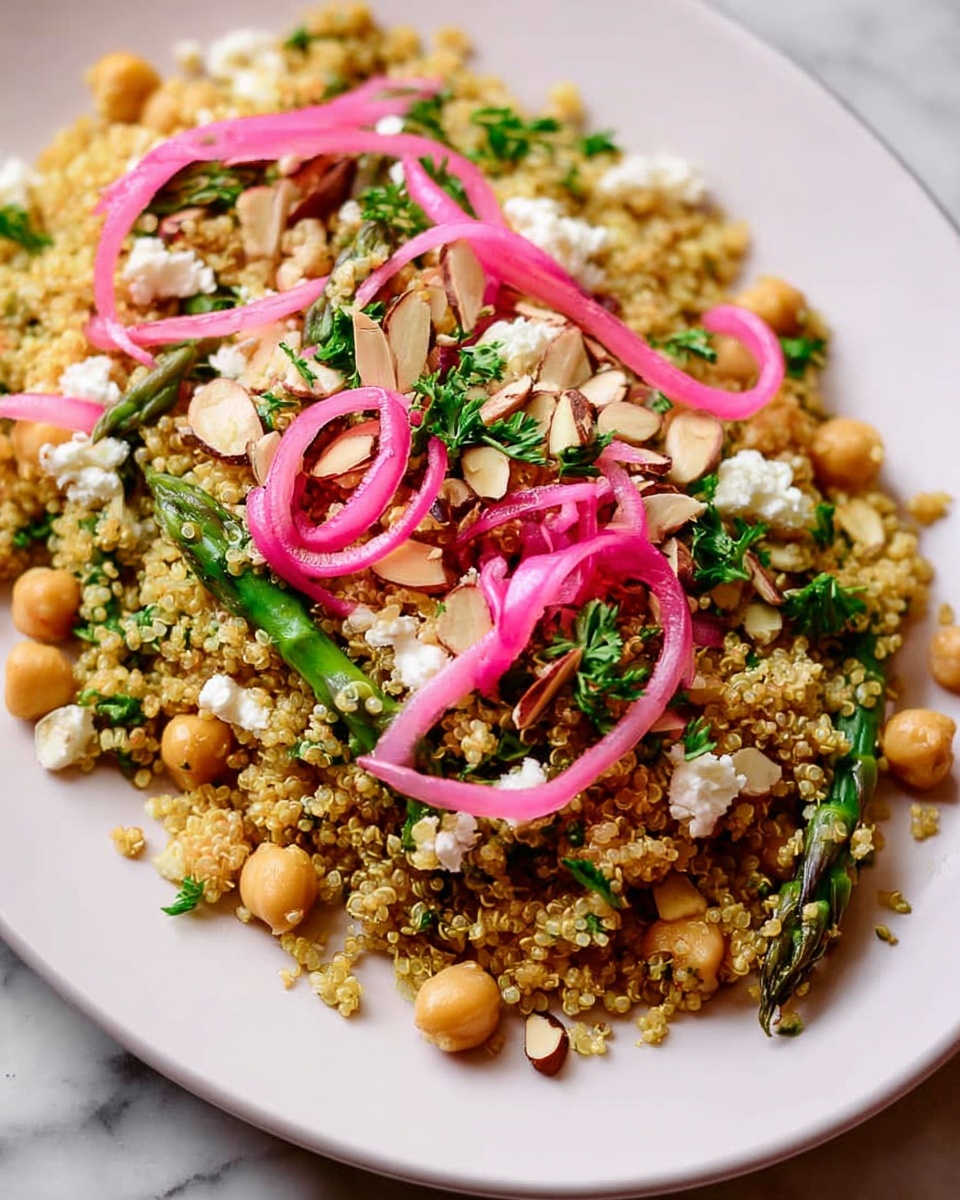 A white plate filled with a layered quinoa salad. The base layer is made of cooked golden quinoa with small grain texture. Intermixed within are light brown chickpeas scattered evenly throughout. There are green pieces of chopped herbs and vegetables like parsley and asparagus adding fresh color and texture. Small white crumbles of cheese are dotted across the dish. On top, bright pink thin spiraled pickled onions are placed in small clusters. Chopped almonds add a crunchy texture and are spread around the salad. The plate is set on a white marbled textured surface. photo taken with an iphone --ar 4:5 --v 7