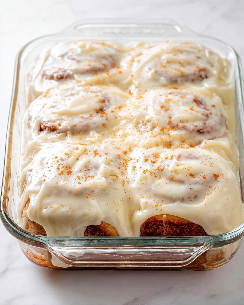 Hawaiian Roll Cinnamon Rolls with Cream Cheese Icing Recipe 5 The image shows a glass baking dish filled with three cinnamon rolls stacked closely together forming one layer. They are covered with a thick, creamy white icing that spreads unevenly across the top, slightly dripping over the edges. The cinnamon rolls have a light brown color visible through the icing, and a few specks of cinnamon powder are sprinkled on top, adding a light orange texture to the white icing. The background has a clean white marbled surface. Photo taken with an iphone --ar 4:5 --v 7