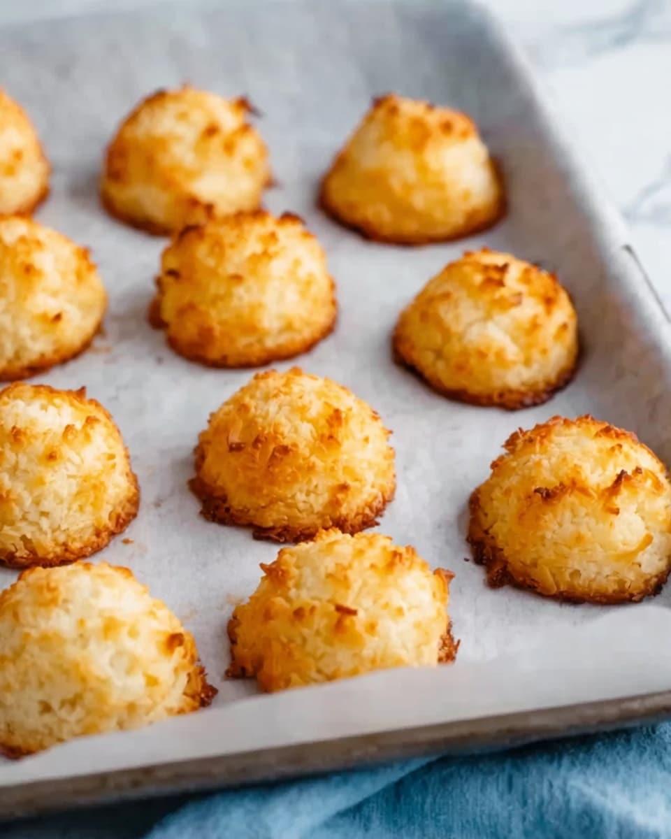 Coconut Macaroons Recipe 5 The image shows a gray rectangular tray filled with round, golden-brown coconut macaroons. Each macaroon has a rough, textured surface from toasted shredded coconut, and they are piled closely together, covering most of the tray. The tray is placed on a green cloth with a white marbled surface underneath. A few small white coconut flakes are scattered around the tray. Photo taken with an iphone --ar 4:5 --v 7