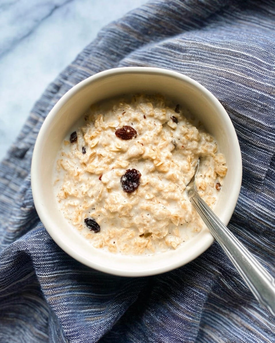 A white bowl filled with creamy oatmeal that has a slightly chunky texture showing oats mixed with milk or cream, and a few visible dark raisins scattered inside. There is a silver spoon placed inside the bowl on the right side. The bowl sits on a dark blue and white striped cloth, all on a white marbled surface. photo taken with an iphone --ar 4:5 --v 7