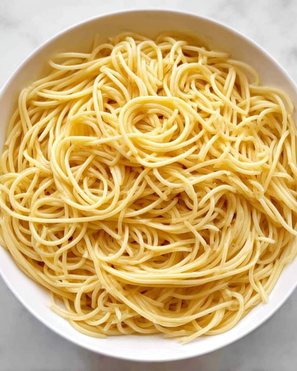 A white bowl filled with plain cooked spaghetti noodles. The noodles are light golden yellow and smooth, tangled loosely with some strands overlapping and curling. The background is a white marbled surface. Photo taken with an iphone --ar 4:5 --v 7