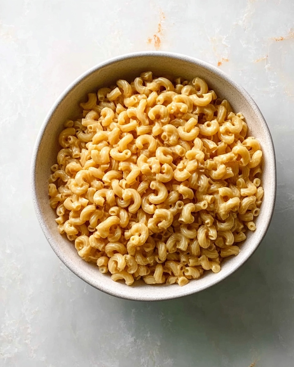 A round white bowl filled with cooked elbow macaroni pasta coated with a light brown sauce. The pasta is evenly mixed and piled to just below the rim of the bowl. The bowl sits on a white marbled surface with subtle grey veins and some faint orange stains around. The lighting is soft and natural, highlighting the texture and slight shine of the macaroni photo taken with an iphone --ar 4:5 --v 7