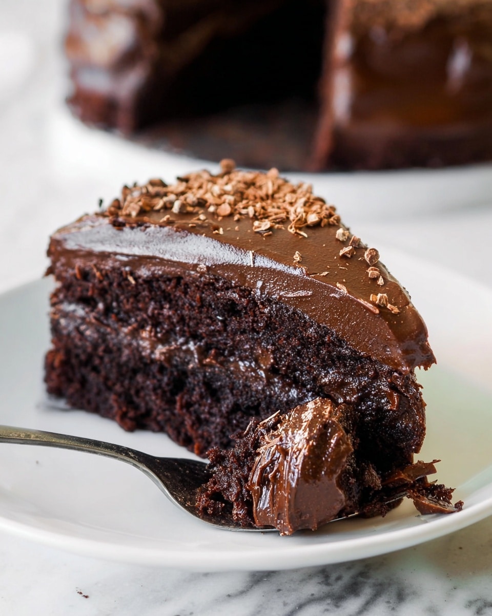 A slice of dark brown chocolate cake with two visible layers sits on a white plate against a white marbled texture background. The bottom layer looks moist and dense, while the top layer is covered with thick, smooth, dark chocolate frosting that shines slightly. On the very top, small chocolate shavings are scattered across the frosting. A silver fork holds a portion of the cake, showing the rich texture and creamy frosting on top. Photo taken with an iphone --ar 4:5 --v 7