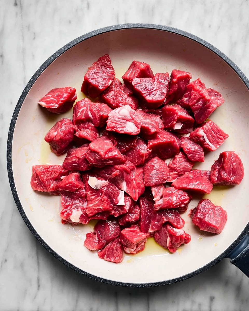 The image shows many small red chunks of raw beef spread across a white pan with a dark edge. Each beef piece has a rich red color with slight white marbling and some fat visible on the surface. The pieces are unevenly spaced with some oil drops shining in the pan, giving a slight wet look. The pan is placed on a white marbled surface. photo taken with an iphone --ar 4:5 --v 7