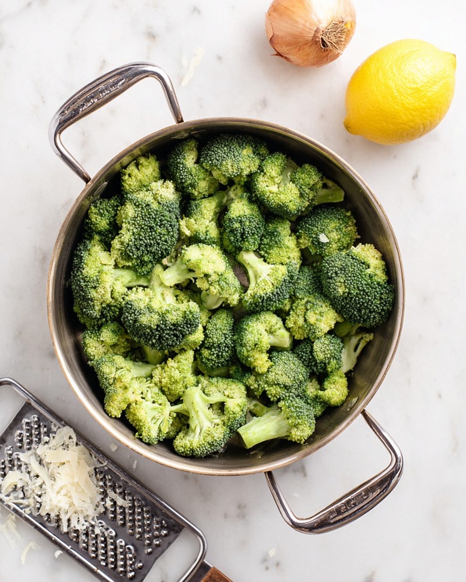 A round silver metal pan with two handles is filled with many green broccoli pieces scattered inside. The broccoli florets are bright green with a rough texture on top and lighter green stems. To the top right of the pan, there is a whole yellow lemon and a half lemon with a brown shallot bulb next to it. At the bottom left corner, a woman’s hand is holding a silver grater with some white cheese shavings around it. The whole scene is set on a white marbled surface. photo taken with an iphone --ar 4:5 --v 7