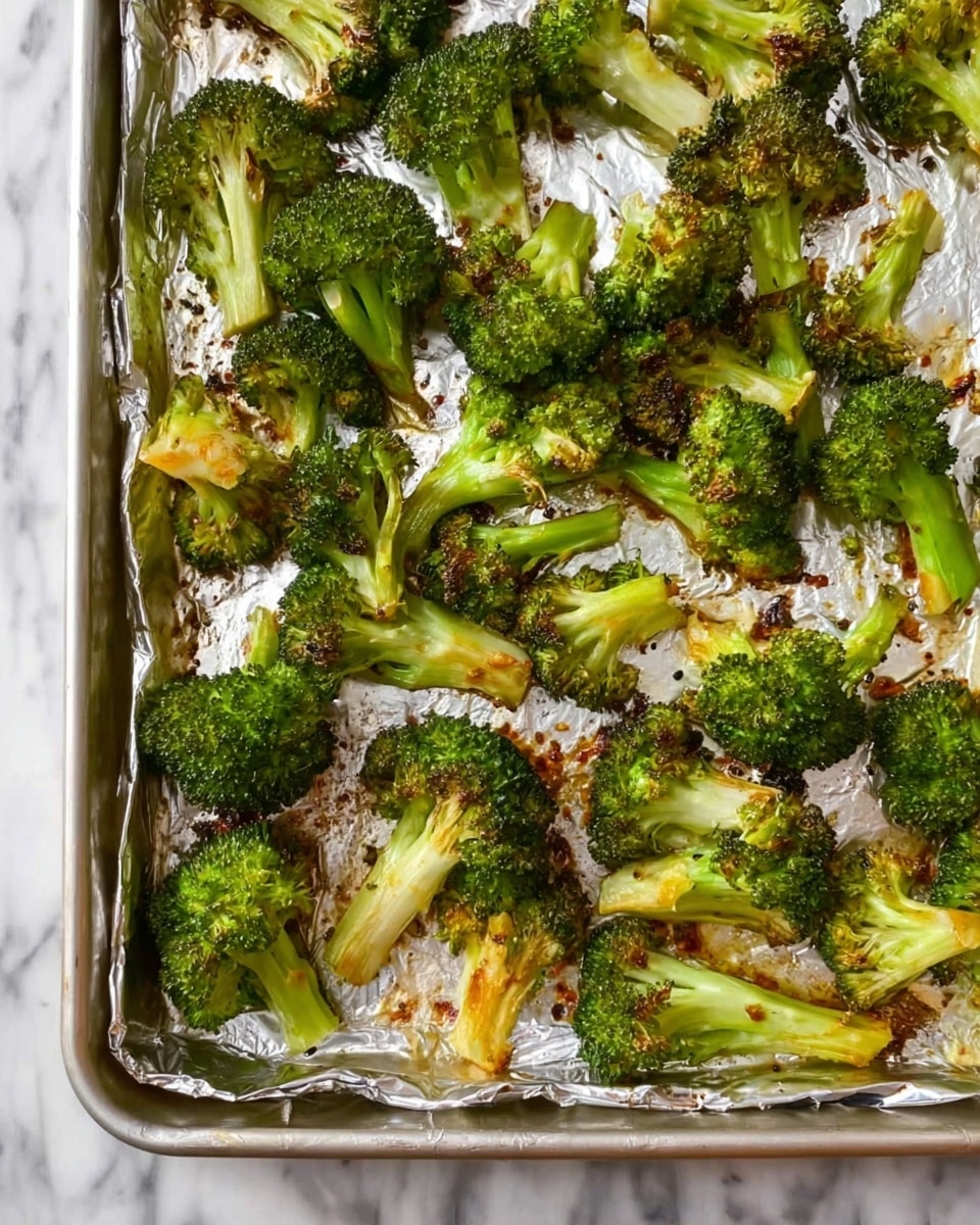 A sheet tray lined with crinkled foil holds many pieces of roasted broccoli, each piece showing green florets with browned and slightly crispy edges, while the white stems underneath are tender and light golden. The broccoli is scattered evenly across the tray, with some parts showing more charred spots for texture and color contrast. The overall look is fresh and simple, with the green and brown colors standing out against the shiny foil and the white marbled surface beneath. Photo taken with an iphone --ar 4:5 --v 7