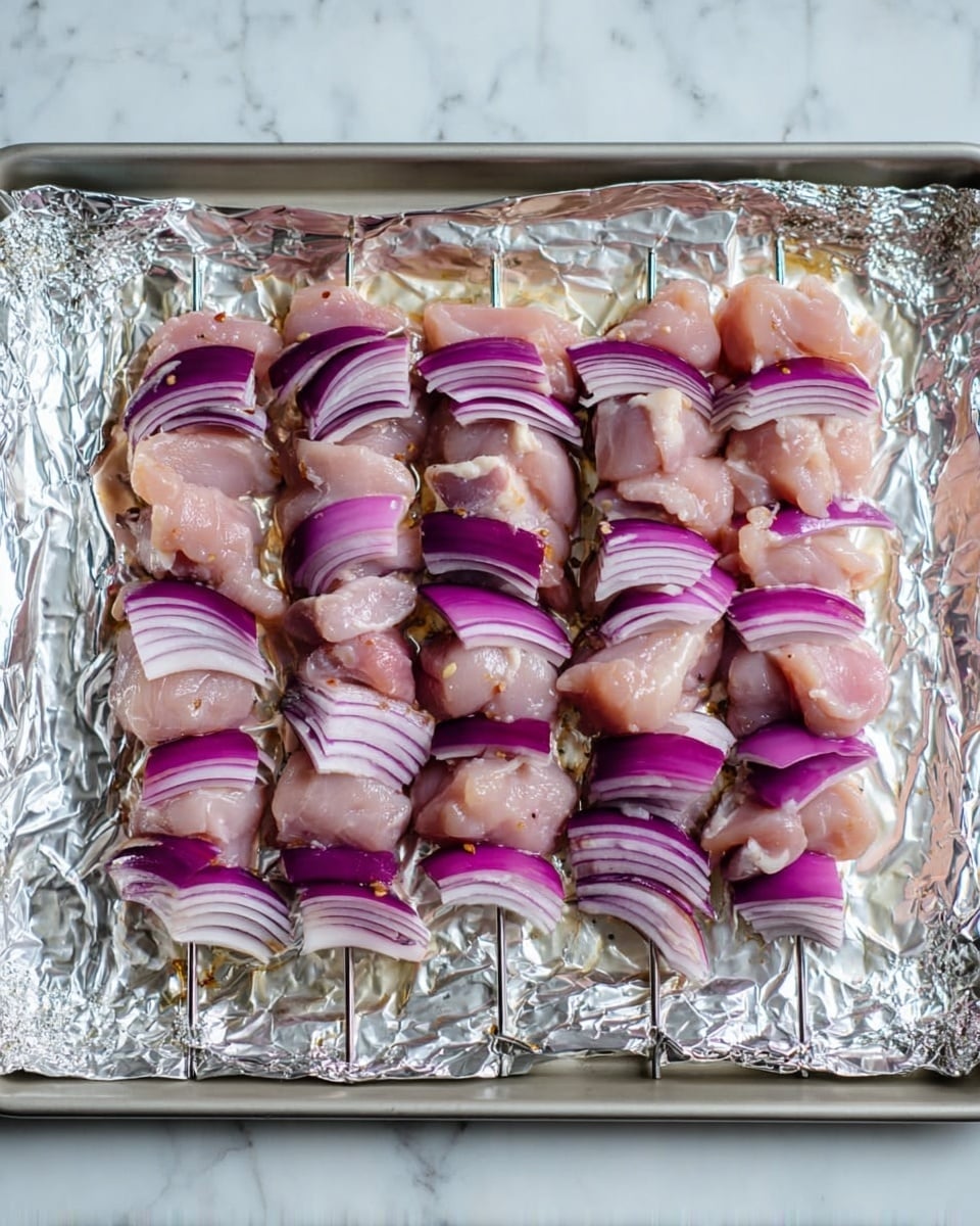 The image shows five metal skewers placed side by side on a foil-lined tray. Each skewer has alternating layers of raw pink chicken pieces and thick slices of purple and white onion, arranged evenly along the length. The foil underneath has a shiny crinkled texture and the tray sits on a white marbled surface. photo taken with an iphone --ar 4:5 --v 7