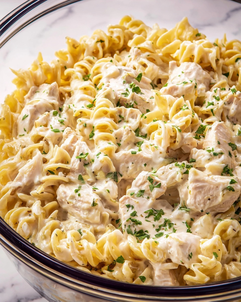A close-up of creamy pasta with many swirled noodles coated evenly in a light yellow sauce. Small green herb pieces are sprinkled across the pasta, adding color contrast. A wooden spoon is lifting a large scoop of the pasta from a pot, showing the sauce's thick, smooth texture. The pot has a dark rim visible in the background, and the white marbled surface underneath sets a clean scene. The image looks fresh and rich in texture, photo taken with an iphone --ar 4:5 --v 7