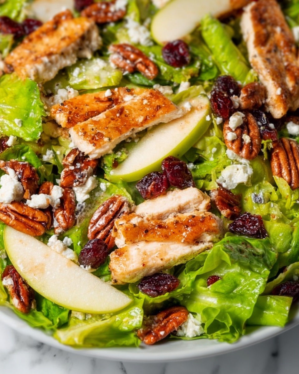 The image shows a close-up of a salad with several layers. The base layer consists of bright green lettuce leaves with a fresh and crisp texture. Scattered on top are pieces of golden-brown cooked chicken strips, which have a slightly crispy outer layer and juicy inside. There are also medium-sized brown pecan nuts spread evenly, adding a smooth and shiny texture. Thin slices of pale green apple are placed around the salad, showing a smooth and moist surface. Small pieces of dark red dried cranberries are visible, adding a chewy texture and rich color contrast. White crumbled cheese is sprinkled lightly over the salad, offering small, soft textures and bright color spots. The salad is on a clean white plate set on a white marbled surface. The photo taken with an iphone --ar 4:5 --v 7