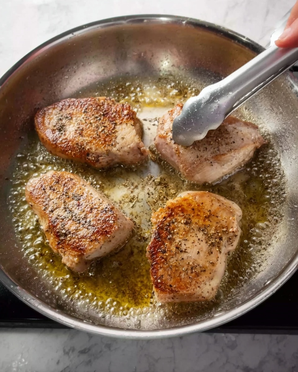 The image shows four pieces of seasoned meat cooking in a shiny stainless steel pan filled with bubbling oil. Three pieces are golden-brown on top with specks of black pepper and spices, while one piece is lighter and less cooked. A woman's hand holding metal tongs is lifting the lighter piece from the pan. The background shows a clean white marbled kitchen surface. photo taken with an iphone --ar 4:5 --v 7