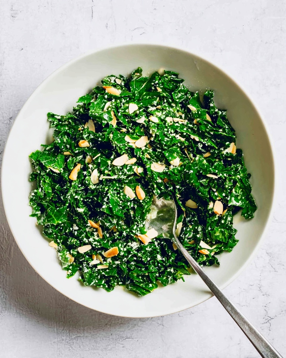 A white bowl filled with finely chopped dark green kale mixed with small pieces of light beige almond slices scattered on top. The texture looks fresh and slightly wet, with a few bits of grated white cheese spread through the kale. A shiny silver spoon is placed inside the bowl, resting on the kale with some kale on the spoon. The bowl is set on a surface with a white marbled texture. photo taken with an iphone --ar 4:5 --v 7