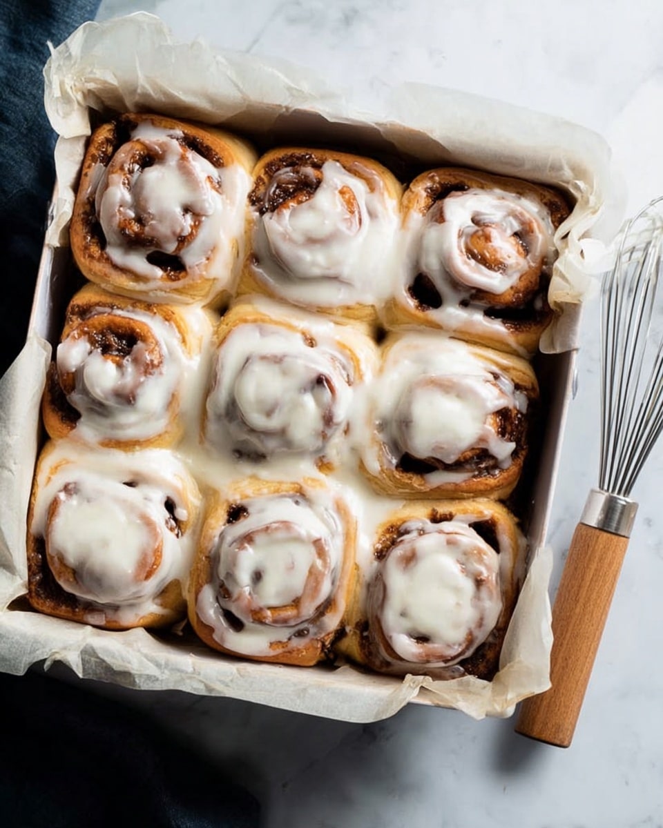 The image shows nine cinnamon rolls arranged in a 3 by 3 pattern inside a square white baking dish lined with parchment paper. Each roll has a swirl pattern with golden brown dough and darker cinnamon filling visible. The top of each roll is generously covered with white creamy icing that is smooth and slightly melted, dripping slightly down the sides. The baking dish sits on a white marbled surface, and a whisk with a wooden handle is placed to the right of the dish. Photo taken with an iphone --ar 4:5 --v 7