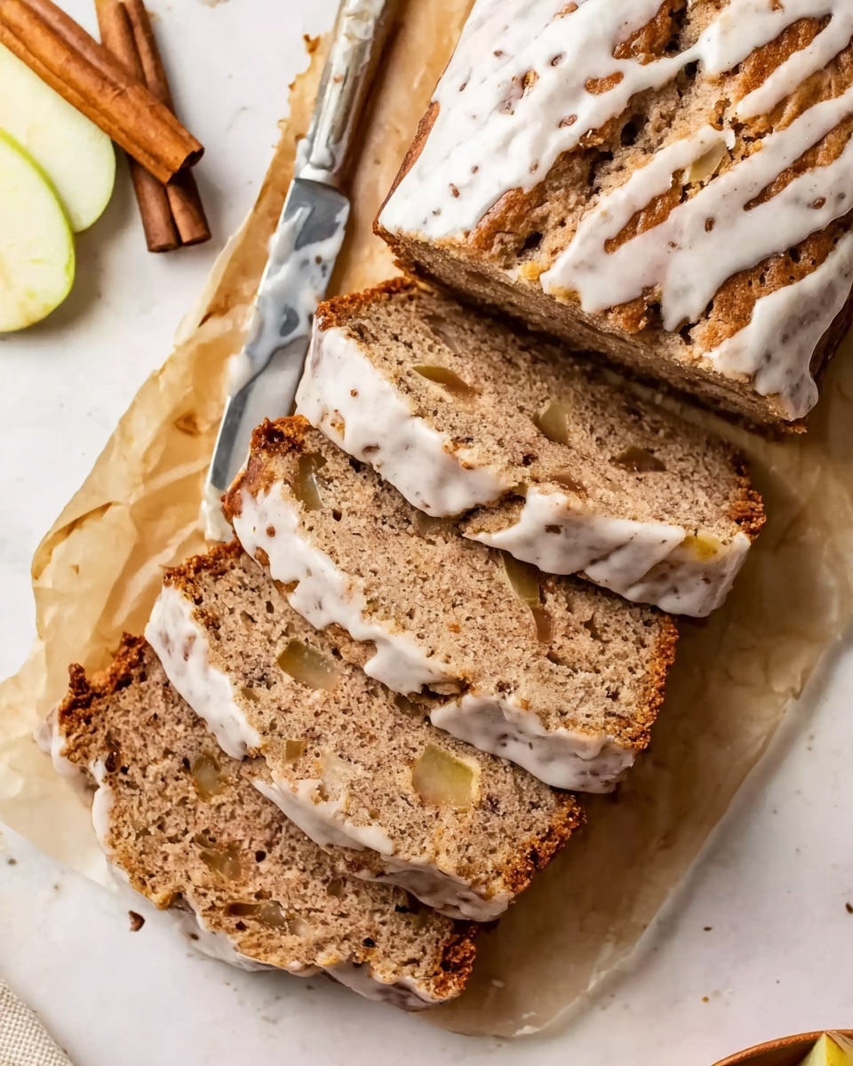 The image shows a white plate covered with parchment paper, holding four slices of spiced bread with pieces of apple inside. The bread has a light brown color with a rough texture dotted with small dark spots and apple chunks. The top of the loaf is drizzled with white icing in uneven lines. Next to the bread is a silver knife resting on the parchment paper, and cinnamon sticks and a slice of apple are placed at the top left corner on the white marbled surface. The lighting highlights the moist texture of the bread and the smooth icing on top, giving a cozy, homemade feel. photo taken with an iphone --ar 4:5 --v 7