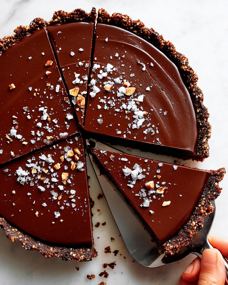 A single slice of chocolate tart on a white plate, showing two layers: a thick, crumbly brown crust with a rough texture at the bottom, and a smooth, shiny dark chocolate layer on top. The chocolate layer is sprinkled with coarse salt flakes, adding contrast to the dark surface. A silver fork lies next to the tart on the right side of the plate. The plate rests on a white marbled surface. Photo taken with an iphone --ar 4:5 --v 7