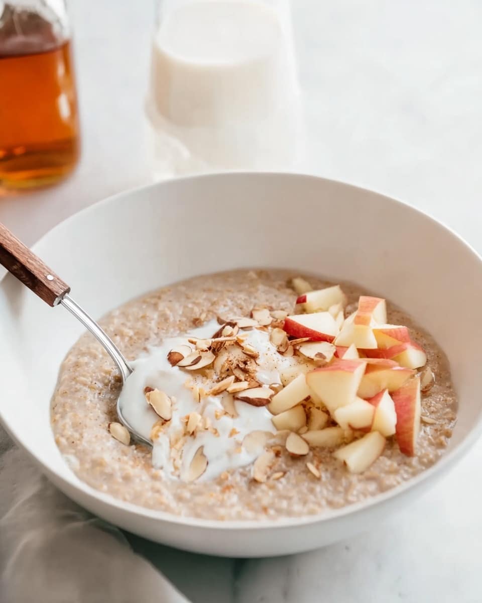 A white bowl holds a creamy, light brown porridge as the base layer, smooth with slight grainy texture. On top, there is a small pile of thin, pale yellow apple strips with red edges arranged in a loose bundle. Over the apple strips, a dollop of thick white yogurt is placed, soft and slightly glossy. To one side of the yogurt, there is a sprinkle of toasted almond slices in warm beige and light brown hues adding a crunchy texture. The bowl sits on a white marbled surface, with a shiny silver spoon resting beside it. Photo taken with an iphone --ar 4:5 --v 7