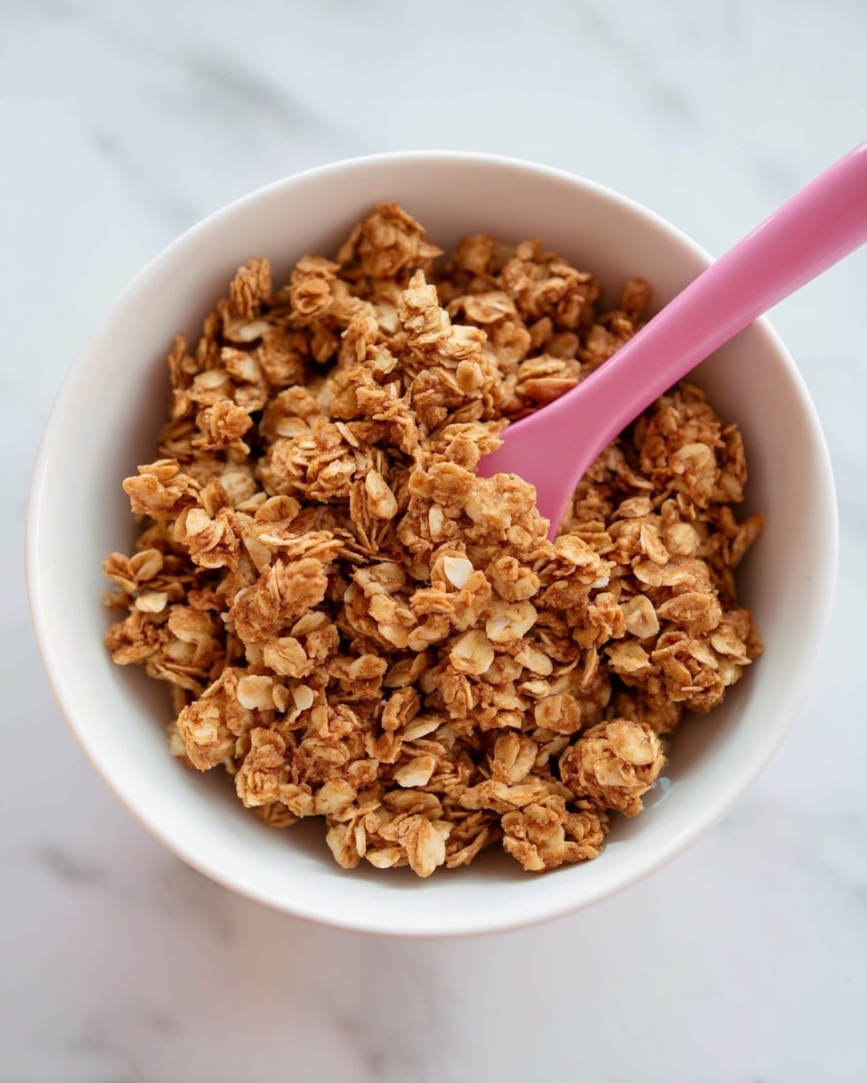 A close-up view of a white bowl filled with light brown granola clusters that have a rough, crumbly texture. A pink spatula with a smooth surface rests inside the bowl, partially covered with the granola mix. The bowl is placed on a white marbled surface, adding a clean and bright background to the scene. The granola pieces vary in size, some showing small bits of oats and grains, giving the mix a natural, hearty look. Photo taken with an iphone --ar 4:5 --v 7