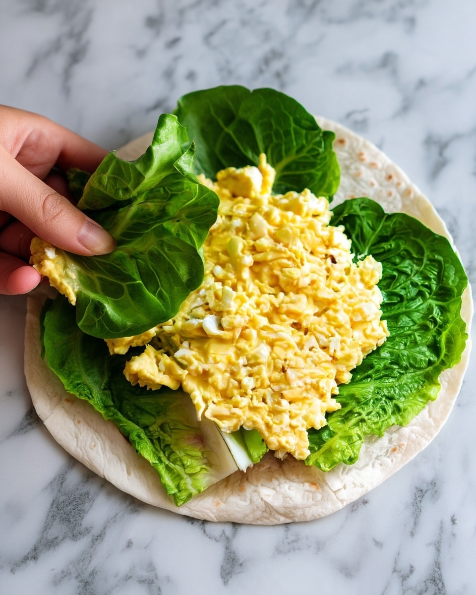 The image shows a soft white tortilla on a white marbled surface with two large green lettuce leaves placed in the center. On top of the lettuce is a thick layer of light yellow egg salad, with small soft chunks and a creamy texture spread evenly. A woman's hand is lifting part of the tortilla from the side. Photo taken with an iphone --ar 4:5 --v 7