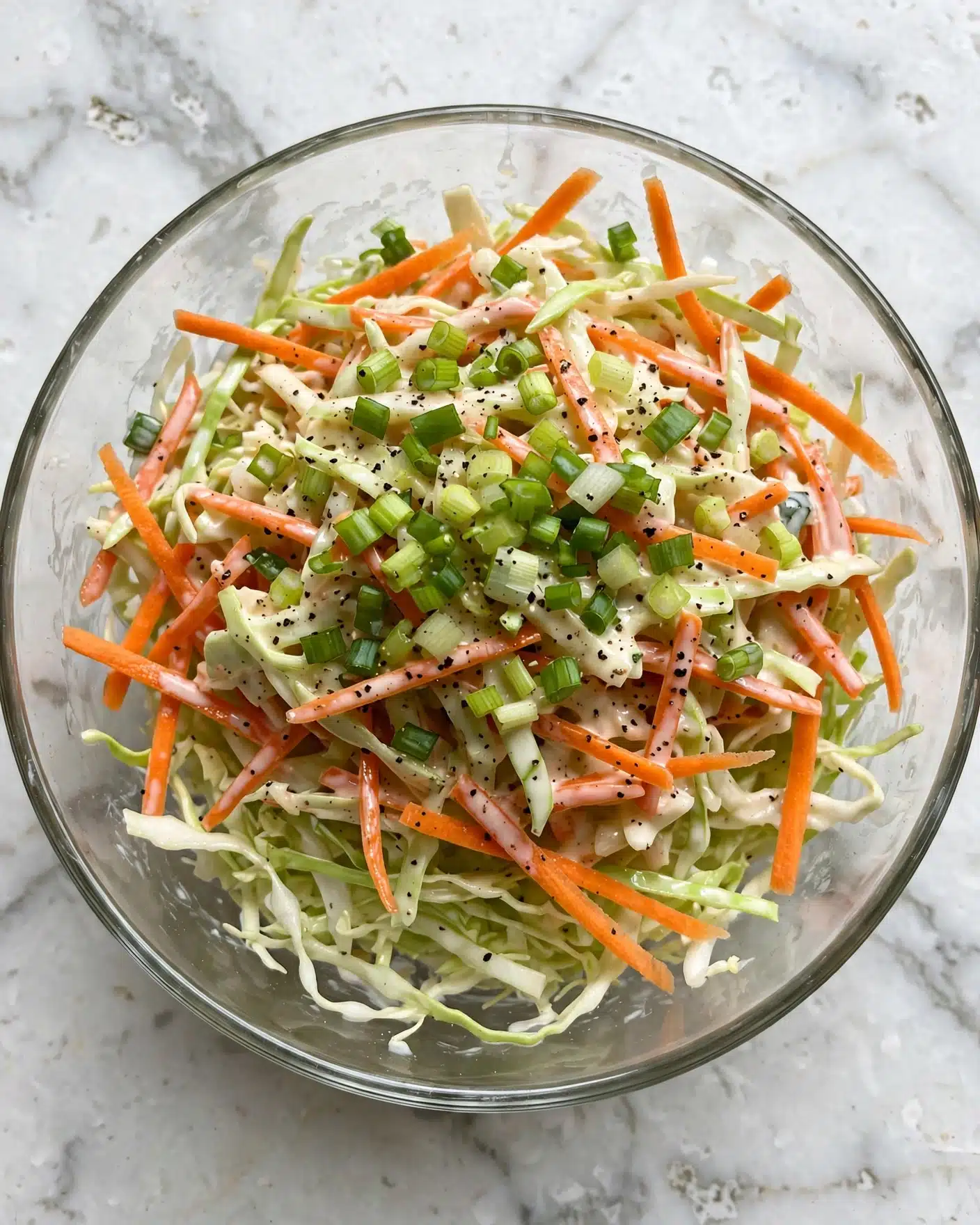 A clear glass bowl filled with a colorful coleslaw mix is shown from above on a white marbled surface. The coleslaw has three main layers: thin, pale green shredded cabbage making the base, bright orange thin carrot strips scattered evenly throughout, and finely chopped light green scallions spread on the top. There is a light, creamy dressing coating the layers, with black pepper sprinkled over everything. The textures vary from crunchy vegetables to smooth dressing. Photo taken with an iphone --ar 4:5 --v 7