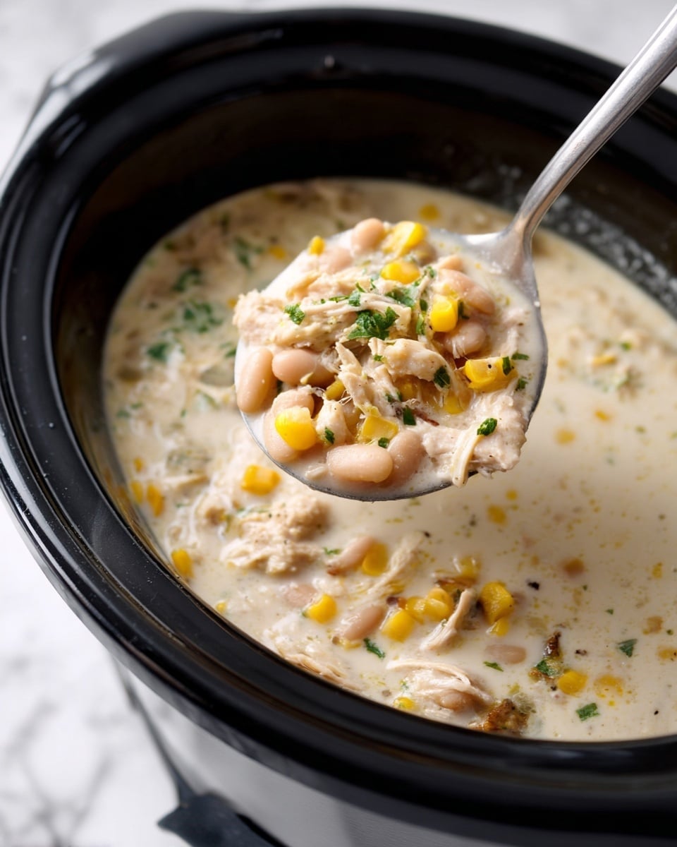 A close-up view of a black slow cooker filled with creamy white soup that has visible chunks of white beans, yellow corn, and shredded light brown chicken mixed throughout. The soup has a thick texture with small green herb pieces sprinkled on top. A silver spoon is lifting a portion of the soup, showing the different ingredients clearly. The slow cooker is placed on a white marbled surface. Photo taken with an iphone --ar 4:5 --v 7
