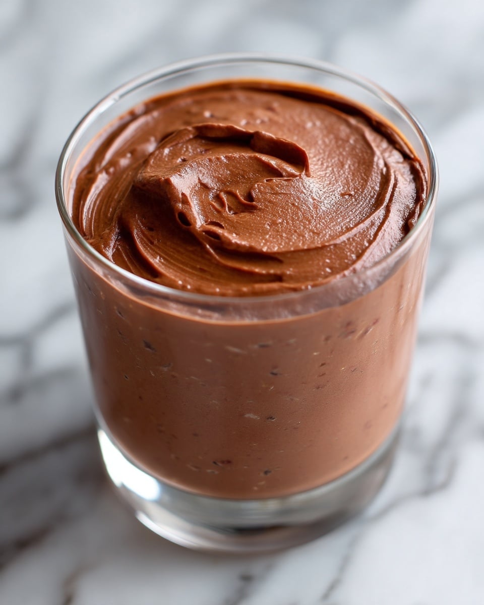 3-Ingredient Low Carb Chocolate Mousse Recipe 5 The image shows a single clear glass filled with smooth chocolate mousse. The mousse has a rich brown color and a creamy, soft texture with gentle swirls on top, giving it a light, airy look. The glass is set on a surface with a white marbled texture. The mousse layer is thick and fills the entire glass, showing hints of darker chocolate bits inside. The photo is sharp and close-up, focusing on the smooth top layer and the glass's round edges photo taken with an iphone --ar 4:5 --v 7