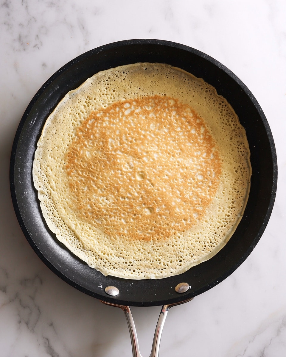 Classic French Crepes with Lemon and Sugar Recipe 4 A thin, light beige pancake is cooking in a black frying pan with a metal handle, spread evenly across the pan's flat surface with some slightly uneven texture and small bubbles near the edges. The pancake has a smooth, soft look with a few creamy white spots where the batter is still wet. The frying pan is set on a white marbled surface, giving a clean and bright background to the scene. photo taken with an iphone --ar 4:5 --v 7