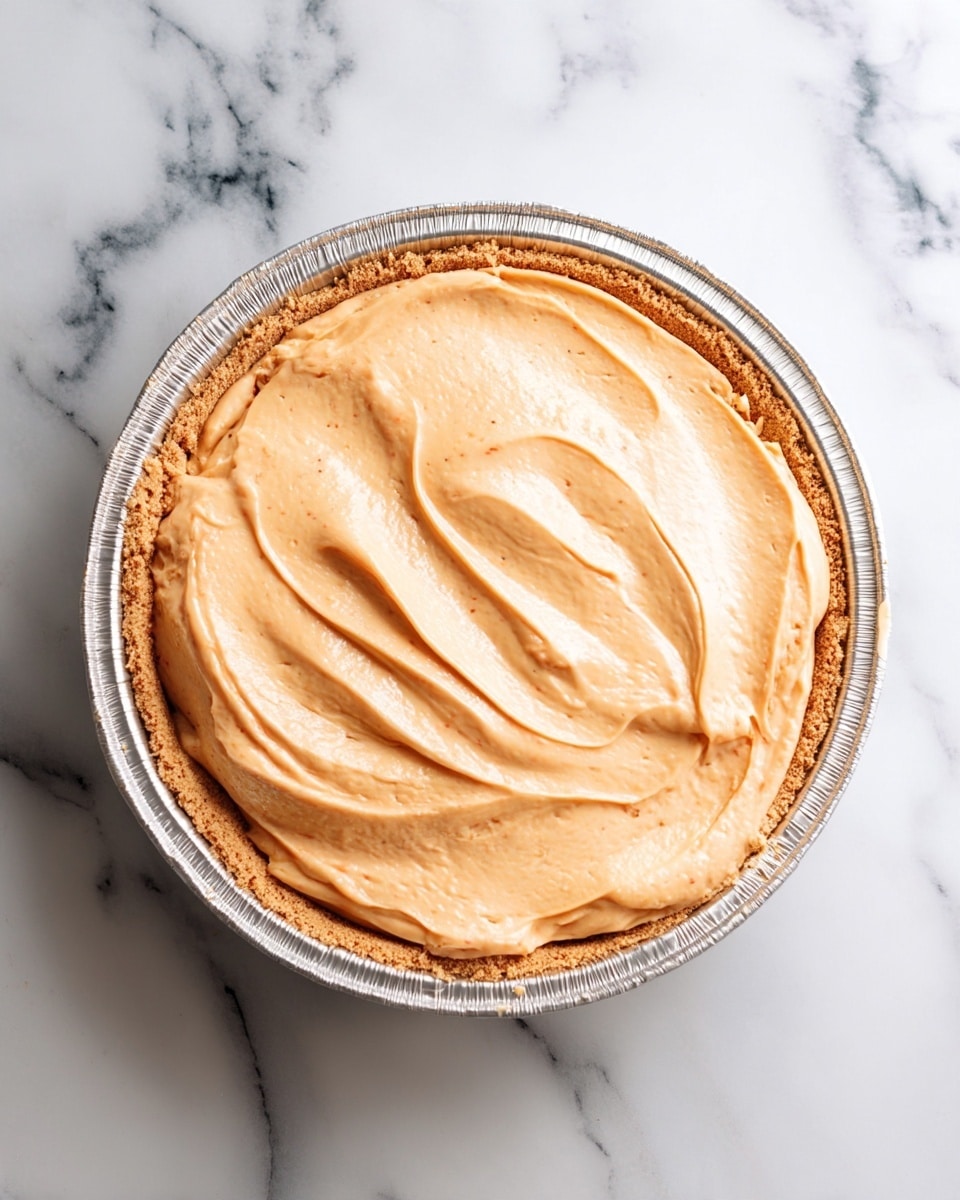 A round pie in a silver foil pan sits on a white marbled surface. The pie has two visible layers: the bottom layer is a light brown crumb crust pressed firmly against the edges of the pan, and the top layer is a thick, smooth, and creamy filling with a pale orange color, spreading evenly to the edges with gentle swirls. photo taken with an iphone --ar 4:5 --v 7