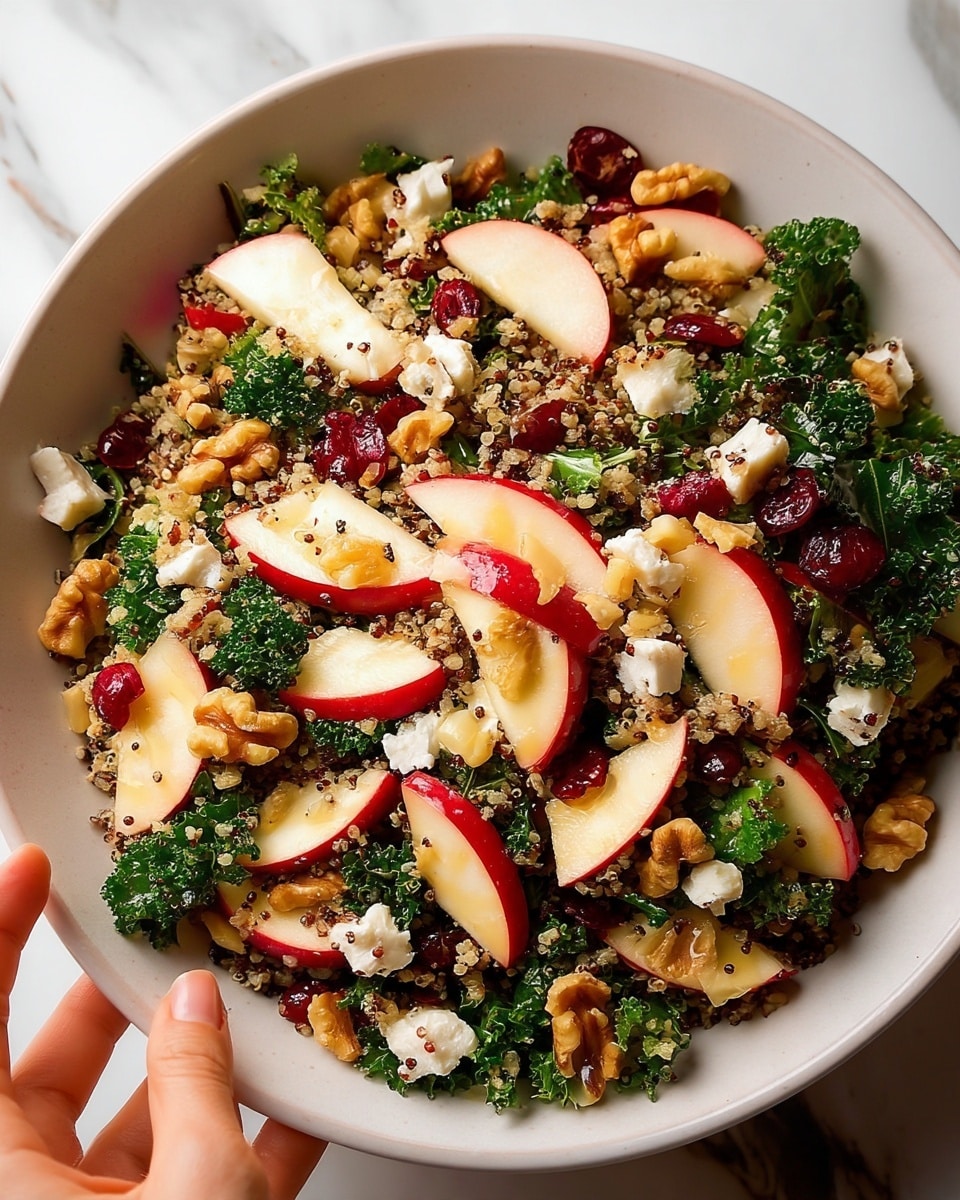 A close-up view of a fresh salad in a large white bowl with ridged sides, filled with layers of finely chopped dark green kale leaves, thin slices of red-skinned apple, small white quinoa grains mixed evenly throughout, and scattered pieces of crumbled white cheese. Bright red dried cranberries and light brown walnut halves are spread on top, adding texture and color contrast. A wooden spoon is partially visible on the right side inside the bowl, all placed on a white marbled surface. photo taken with an iphone --ar 4:5 --v 7