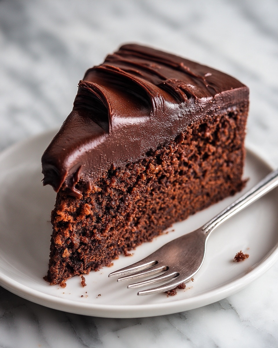 Easy Chocolate Fudge Cake Recipe 5 The image shows a close-up of a single slice of chocolate cake on a white plate with a white marbled texture background. The cake has two main layers: a dense, moist-looking dark brown cake layer with a slightly crumbly texture and a thick, smooth dark chocolate frosting layer on top. The frosting has visible swirls and ridges, giving it a rich, creamy appearance. Some crumbs are scattered near the slice, and a silver fork is placed next to the cake on the plate. Photo taken with an iphone --ar 4:5 --v 7