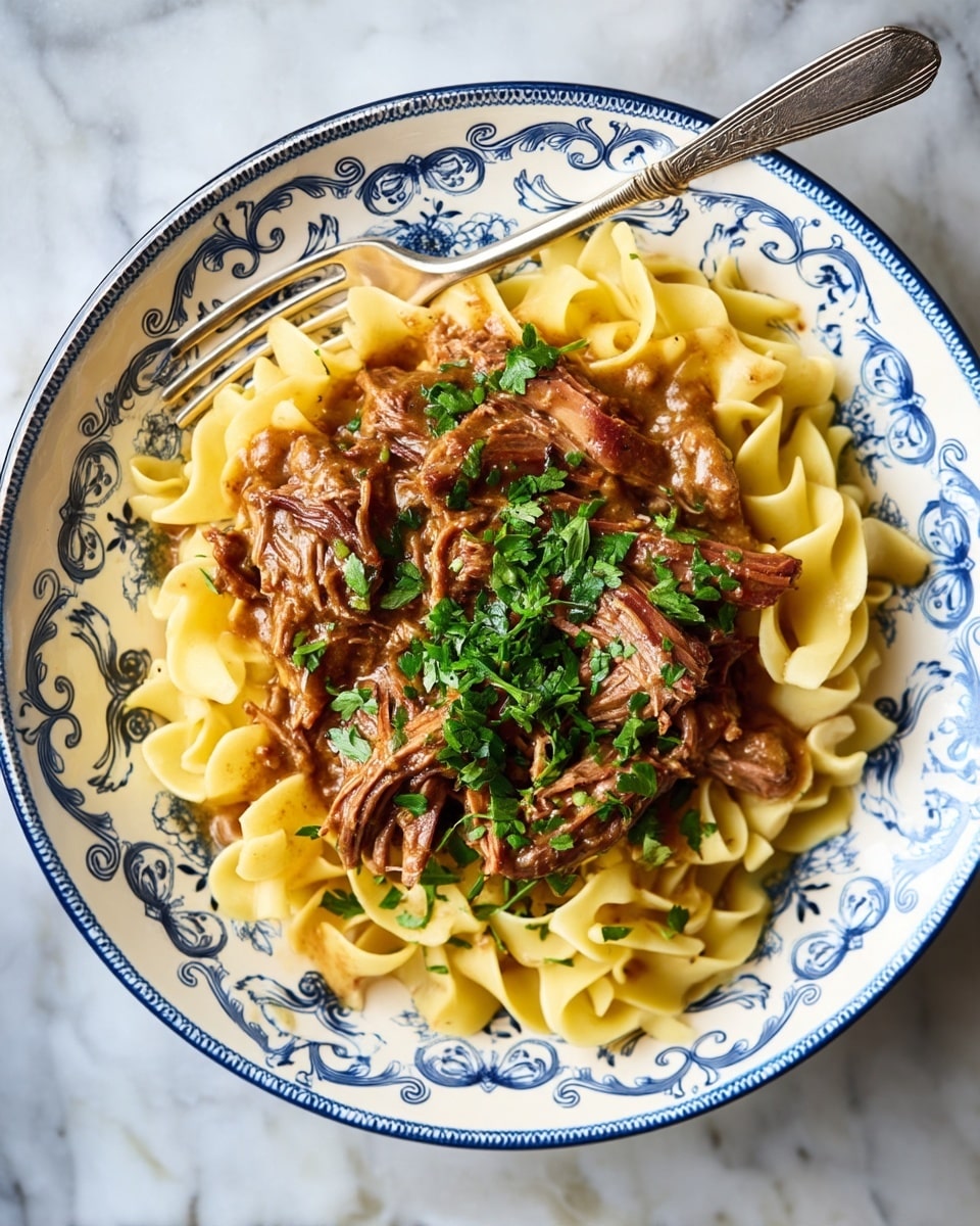 The dish shows a white plate with blue floral patterns filled with three main layers: the bottom layer has pale yellow egg noodles with a soft, slightly curled texture, the middle layer is thick brown shredded meat mixed with a rich gravy sauce, and the top layer is fresh chopped green herbs sprinkled over the meat. A silver fork rests on the edge of the plate against the noodles. The plate is set on a white marbled surface. photo taken with an iphone --ar 4:5 --v 7