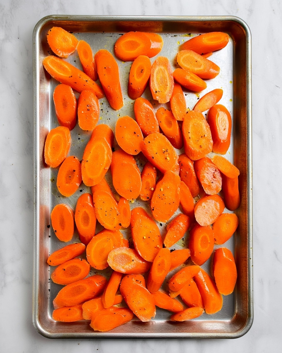 A large silver baking tray holds many slices of bright orange carrots scattered in a single layer, each piece cut into thick, uneven oval shapes with a smooth shiny texture. The carrots are seasoned with tiny black specks of pepper and a light coating of oil that gives them a slight glisten. The tray is set on a white marbled surface, showing some small oil spots and slight dark marks on the tray edges from use. The colors are mainly orange from the carrots and grayish silver from the tray, with a simple, clean setting. photo taken with an iphone --ar 4:5 --v 7