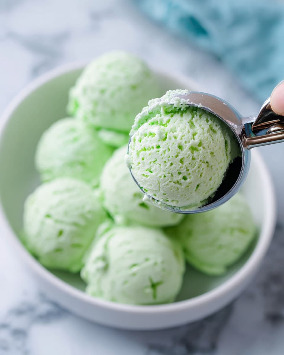 Zombie Punch for Kids Recipe 4 A close-up of a silver ice cream scoop holding a single round scoop of light green ice cream with a creamy texture. In the background, there are several more scoops of the same light green ice cream placed in a white bowl, which sits on a white marbled surface. The ice cream has a soft, slightly fluffy appearance with small uneven bits inside. The background shows an out-of-focus light blue and green area with reflections. photo taken with an iphone --ar 4:5 --v 7