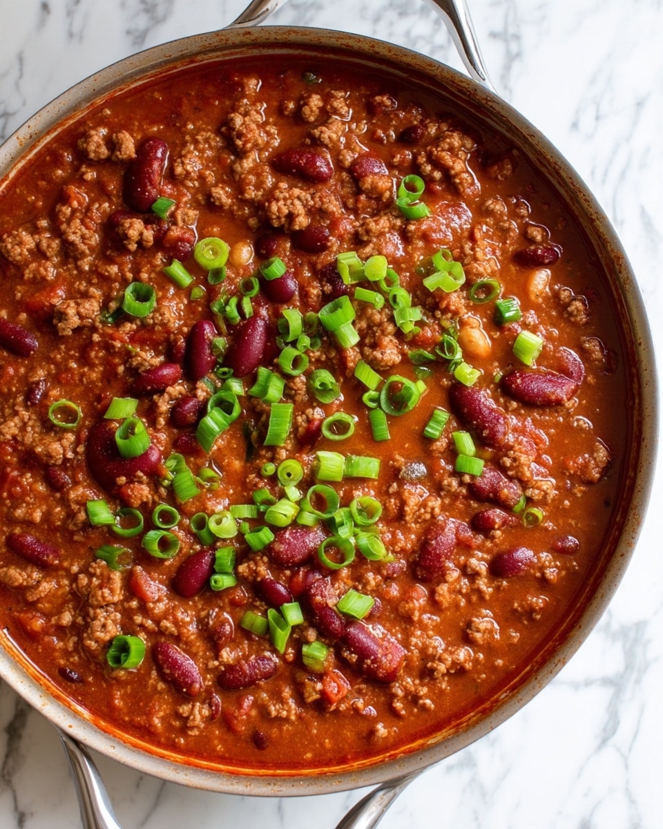 The image shows a close-up view of a thick stew inside a pan, filled with a rich reddish-brown sauce mixed with small chunks of ground meat and whole kidney beans. On top, bright green sliced scallions are scattered evenly, adding a pop of color. The stew's texture looks creamy and smooth with visible bits of meat and beans, giving it a hearty appearance. The pan sits on a white marbled surface. photo taken with an iphone --ar 4:5 --v 7