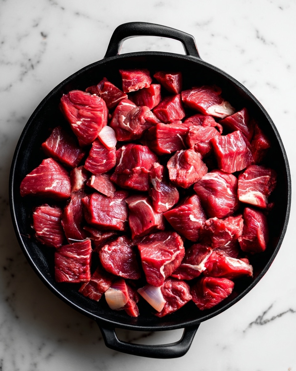 The Easiest Garlic Butter Steak Bites Recipe 4 The image shows many small pieces of raw red meat placed closely together in one layer inside a black cast iron pan. The meat pieces have different shades of red and some light fat parts. The pan is full with the meat pieces, covering all black inner surface, and the texture of the meat looks fresh and moist. The background surface is a white marbled texture. photo taken with an iphone --ar 4:5 --v 7
