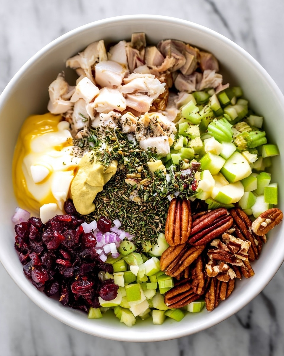 Turkey Salad with Cranberries, Apples, and Pecans Recipe 1 A close-up view of a white bowl filled with layers of ingredients arranged separately: chopped pale cooked chicken pieces at the top, creamy light yellow mayo and mustard beside it, a pile of mixed green herbs and spices sprinkled in the middle, small chopped light green celery and apple pieces on the right side, chopped purple-red onions and dark red dried cranberries in the lower right, and crunchy brown pecan nuts near the center bottom. The bowl sits on a white marbled surface. photo taken with an iphone --ar 4:5 --v 7