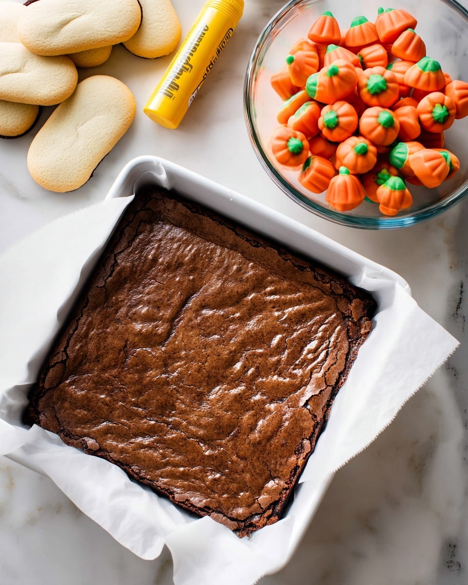 A square white baking dish lined with white parchment paper holds a rich, dark brown baked brownie base with a slightly rough and cracked texture. On the white marbled surface next to it, there are small bright orange pumpkin-shaped candies with green tops, a tube of yellow writing gel, and a stack of oval, light beige cookies with a smooth surface. Also visible is a clear glass bowl filled with a thick, swirled, pale green frosting. Photo taken with an iphone --ar 4:5 --v 7